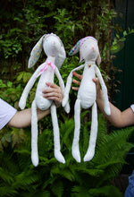 Load image into Gallery viewer, Two handmade rabbit dolls made of white fabric, with patterned ears and bodies, being held up by two children.