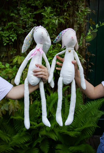 Two handmade rabbit dolls made of white fabric, with patterned ears and bodies, being held up by two children.