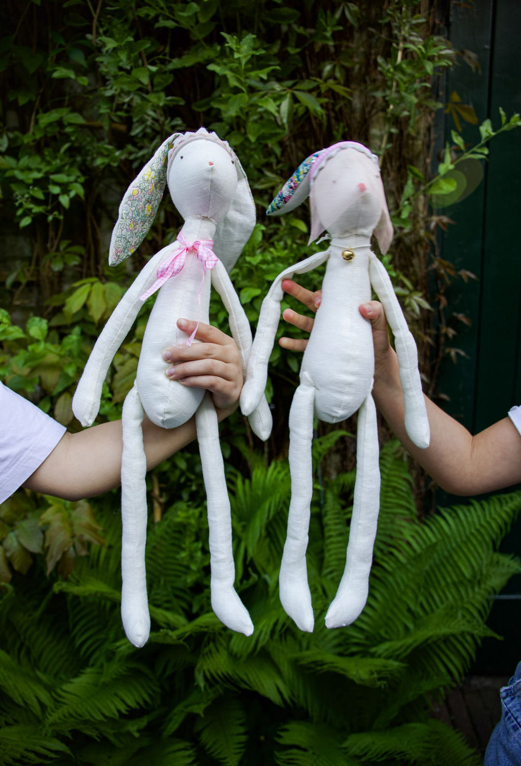 Two handmade rabbit dolls made of white fabric, with patterned ears and bodies, being held up by two children.