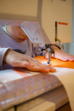 Load image into Gallery viewer, A child is participating in a sewing workshop, sitting at a sewing machine with their hands on the fabric.

